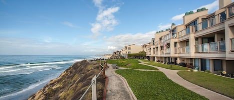 A coastal hotel with a pathway leading to the beach, featuring balconies and a view of the ocean.