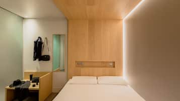 A modern hotel room with a wooden headboard, a desk with a chair, a mirror, and a bed with white linens.