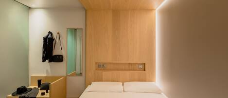 A modern hotel room with a wooden headboard, a desk with a chair, a mirror, and a bed with white linens.