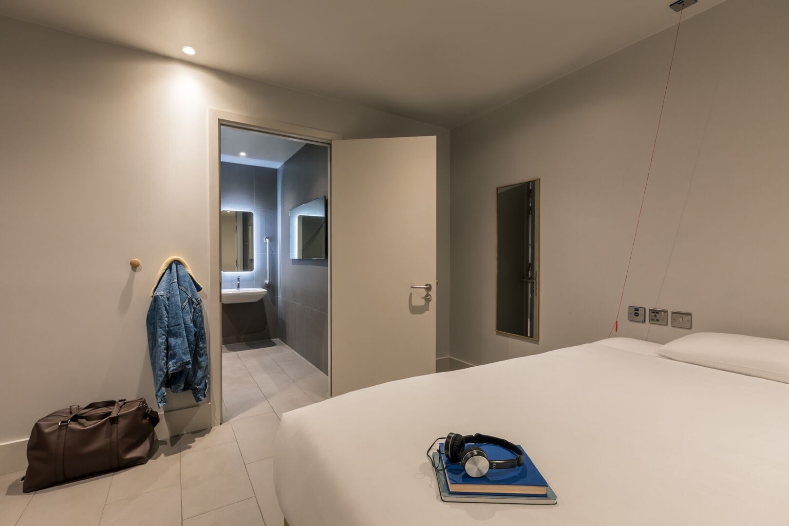 A modern hotel room with a large bed, a bedside table with a book and headphones, a suitcase, and a jacket hanging on a hook.
