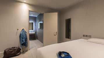 Una habitación de hotel moderna con una cama grande, una mesita de noche con un libro y auriculares, una maleta y una chaqueta colgada de un perchero.