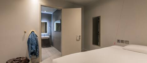 Una habitación de hotel moderna con una cama grande, una mesita de noche con un libro y auriculares, una maleta y una chaqueta colgada de un perchero.