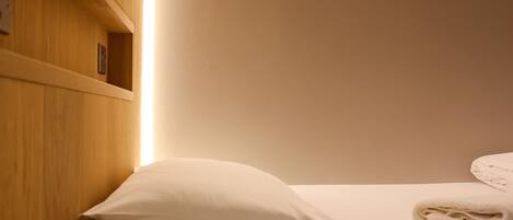 A neatly made bed with a white pillow and folded towels, wooden headboard, and a shelf with a small item on it.