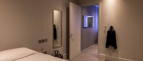 A modern hotel room with a large bed, a mirror, a coat rack, and a bathroom visible through an open door.