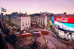 Eine belebte Stadtkreuzung mit historischen Gebäuden, einer großen Coca-Cola-Werbetafel und doppelstöckigen Bussen.