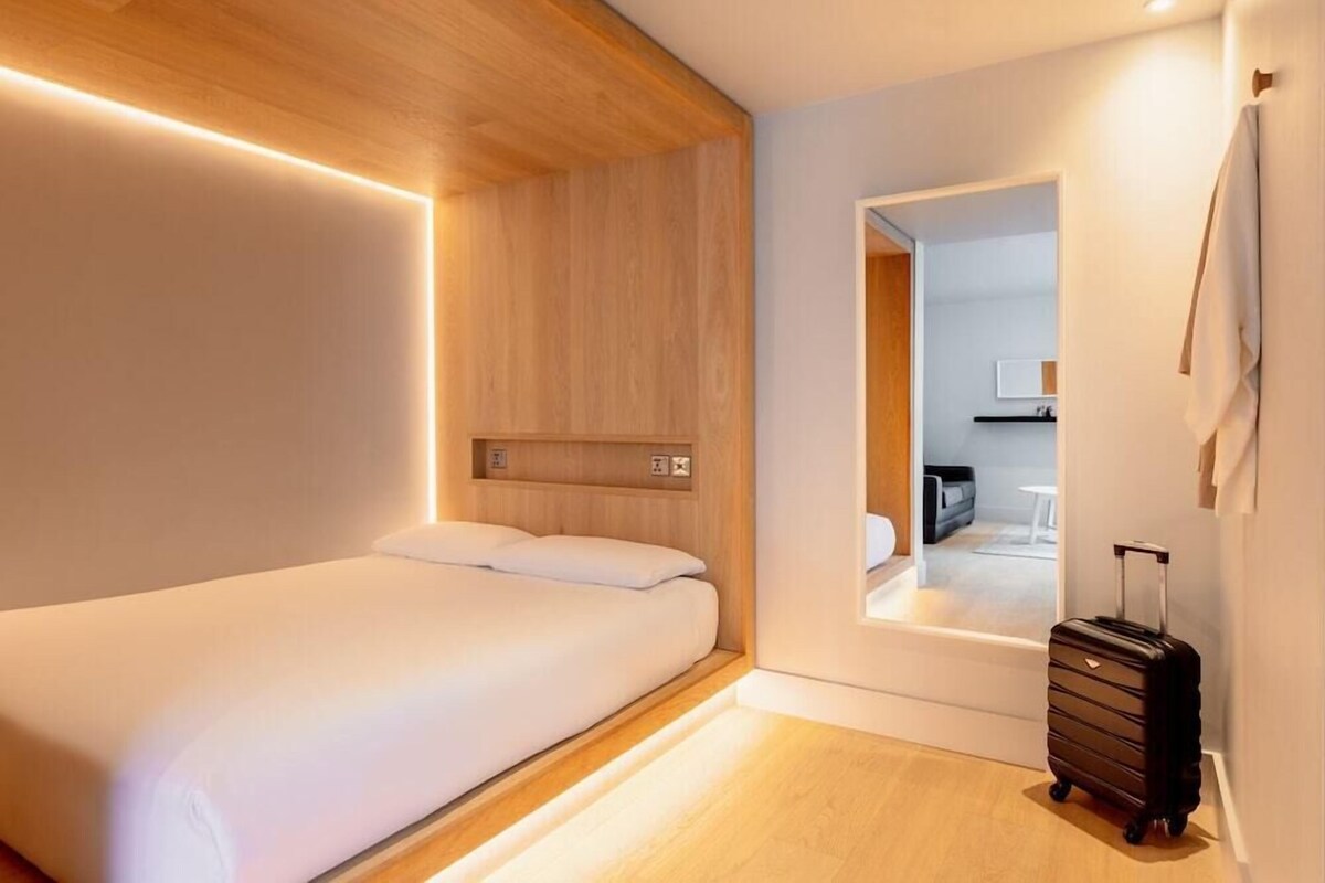 A modern hotel room with a wooden ceiling, a bed, a wall-mounted control panel, and a suitcase.