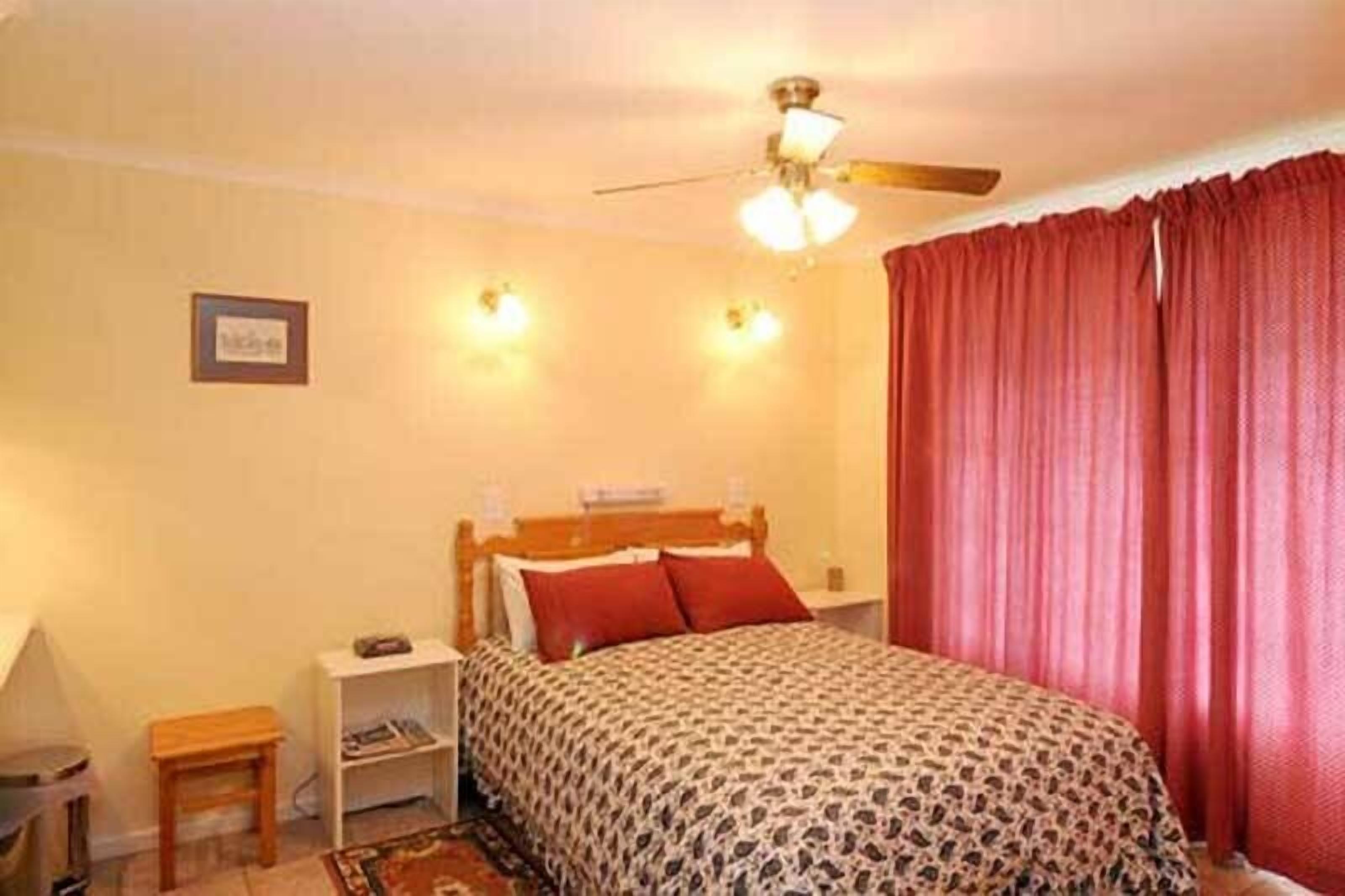 Un dormitorio con cama, mesita de noche, una pequeña mesa, ventilador de techo y cortina roja.