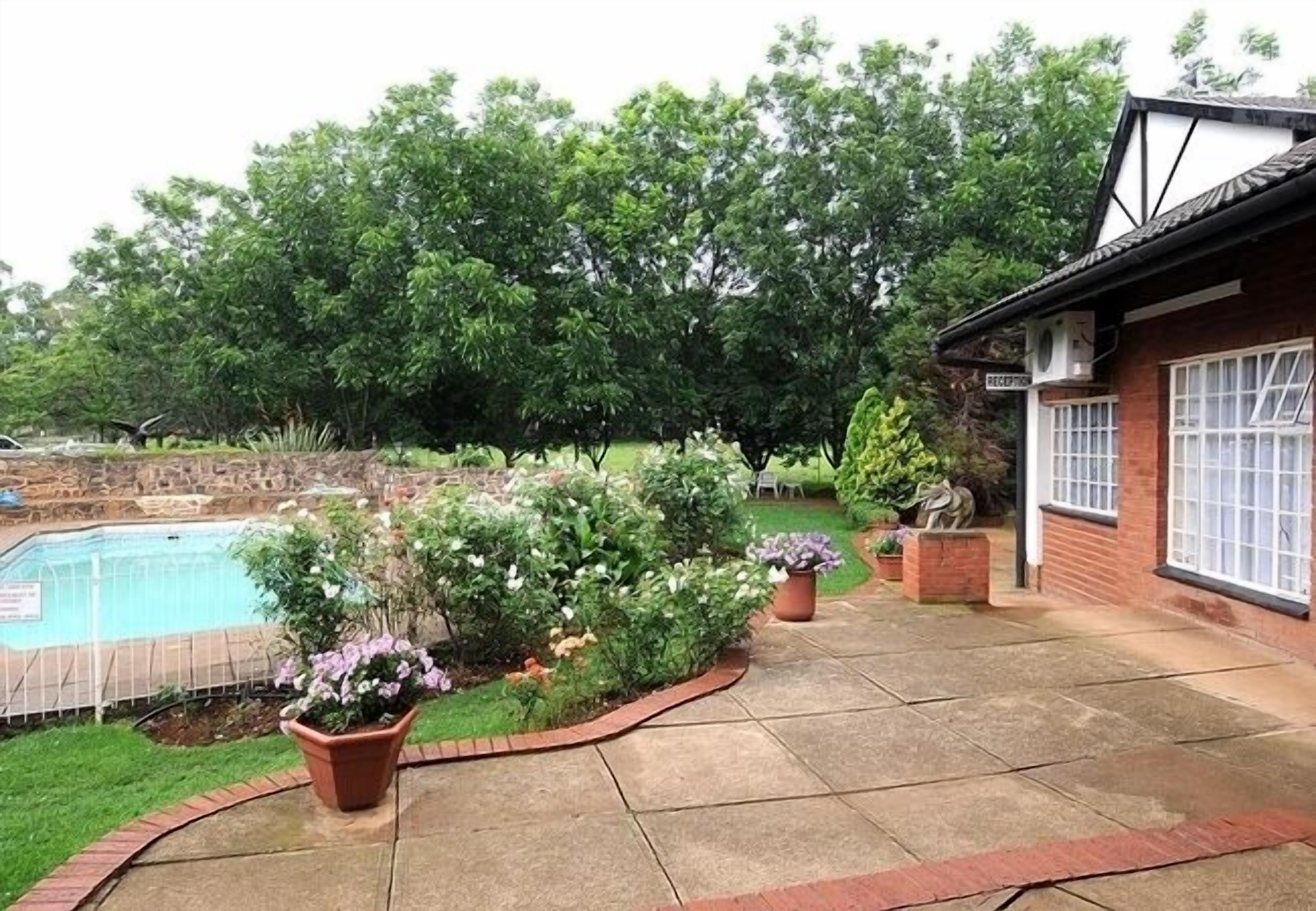A backyard with a swimming pool, a brick house, and a garden with various plants and a statue.