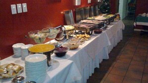 Disposição de buffet com diversos alimentos e utensílios de servir sobre uma mesa coberta por um pano de mesa branco.