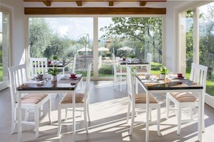 Un comedor luminoso y soleado con sillas y mesas blancas, y ventanas amplias que ofrecen vista al jardín y a los árboles.