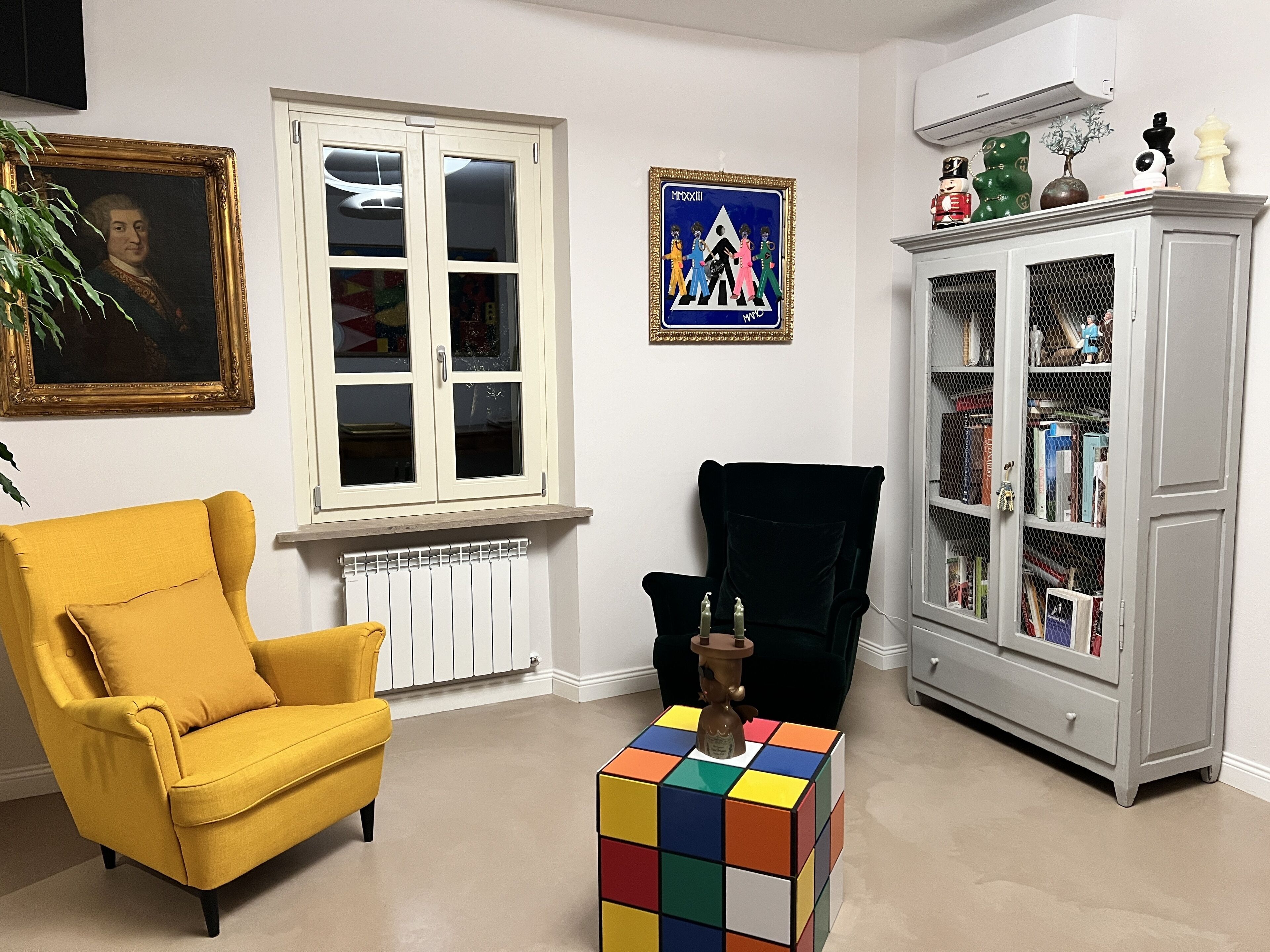 Una habitación con un sillón amarillo, una silla negra, una estantería, un radiador y un cuadro en la pared.