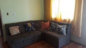 A dark brown sectional sofa with three decorative pillows in a room with light green walls and a window with yellow curtains. - SEASON IN MONGAGUÁ (Balneario Itaguaí)