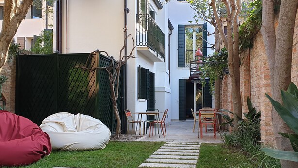 A narrow courtyard with a paved path leading to a white building with black shutters. There are two chairs and a table in the center, surrounded by greenery and a tree. - Casa Giotto (Padova)