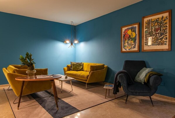 Una sala de estar con pared azul, sofá amarillo, sillón negro, mesa de centro de madera y dos obras de arte enmarcadas.