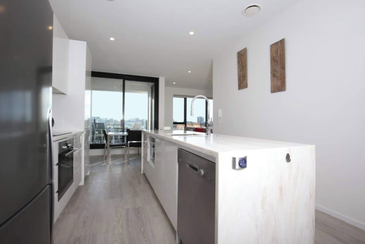 A modern kitchen with a built-in dishwasher, a large island with a sink, and a view of the city through large windows.