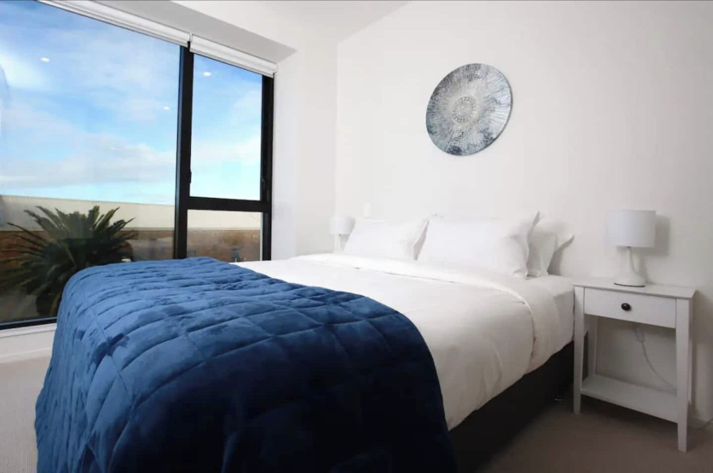 A bedroom with a large bed, a nightstand, a wall-mounted lamp, and a window with a view of the sky and palm tree.