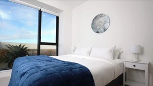 A bedroom with a large bed, a nightstand, a wall-mounted lamp, and a window with a view of the sky and palm tree.