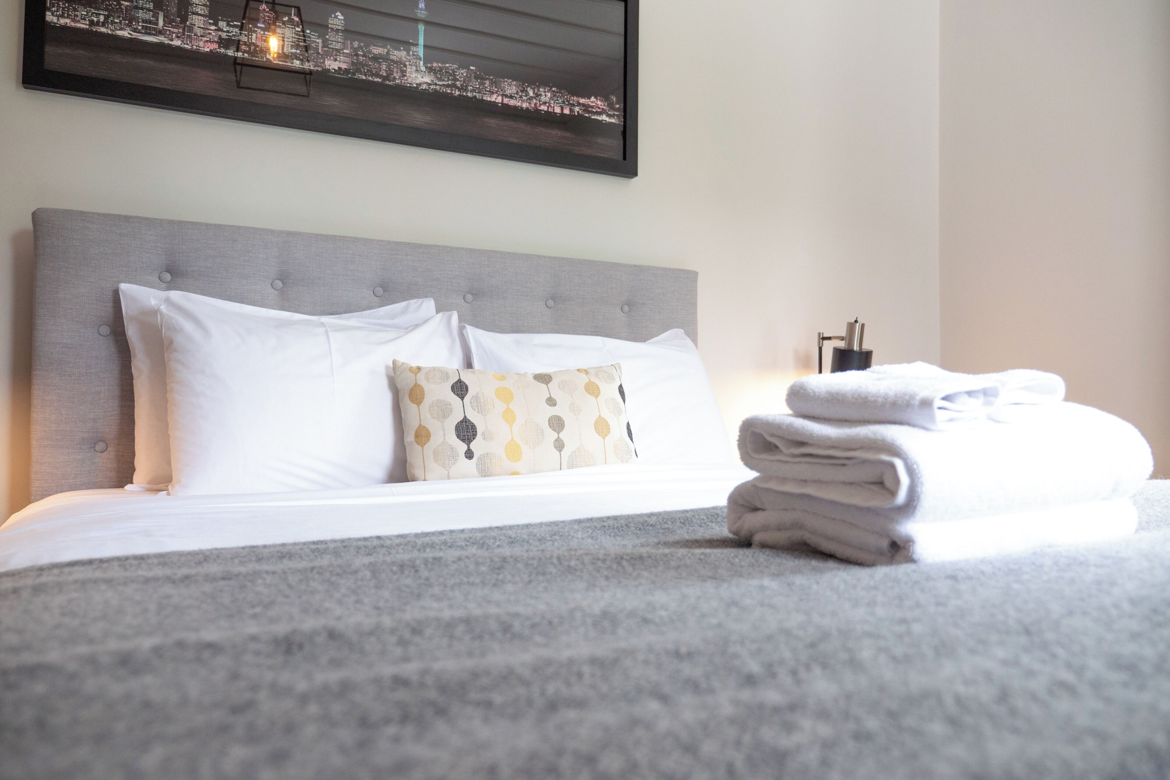 A neatly made bed with white linens and a patterned pillow, a folded towel on the bed, and a cityscape view on the wall.