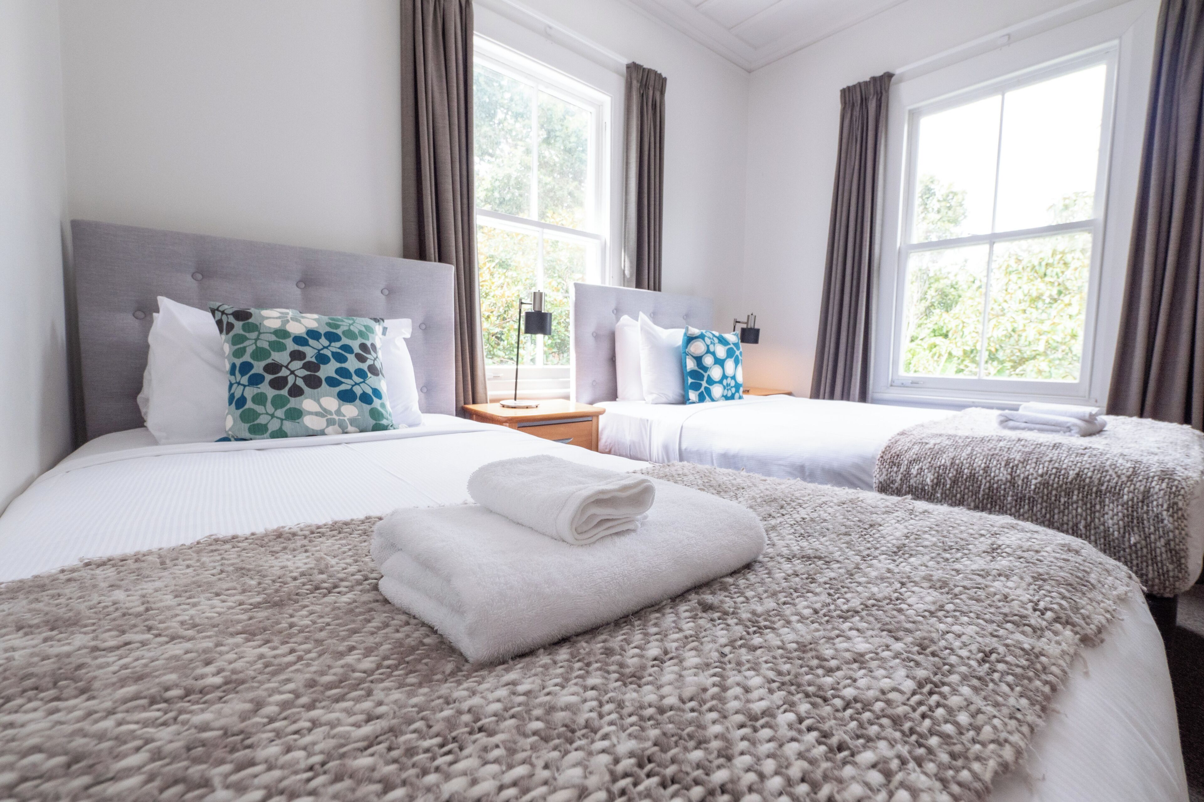 Une chambre d’hôtel avec deux lits, chacun doté d’une tête de lit grise et de draps blancs. Il y a deux fenêtres avec des rideaux, ainsi qu’une table de chevet avec une lampe.