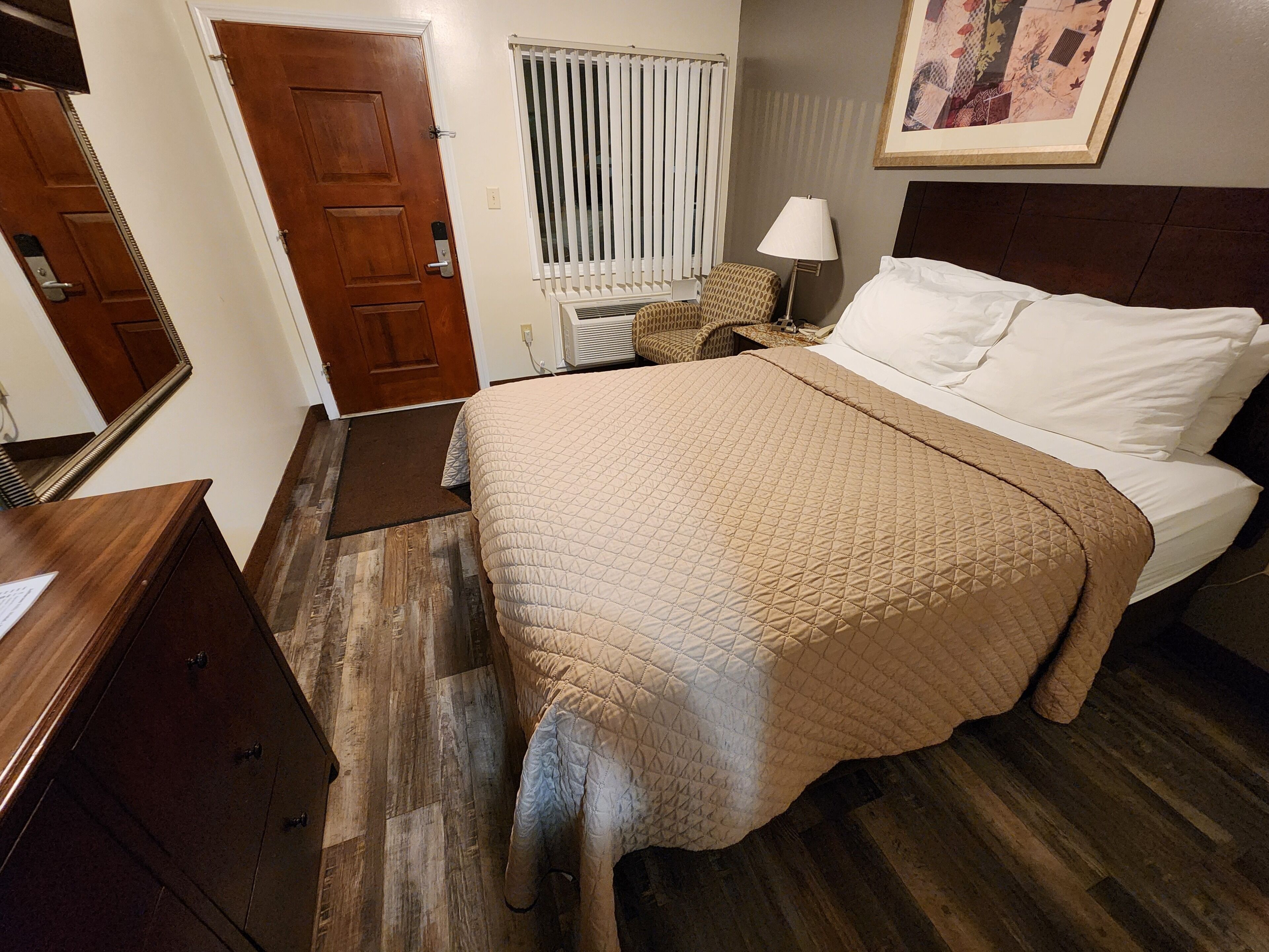 A bedroom with a bed, a nightstand, a lamp, a wooden dresser, a door, and a window with blinds.
