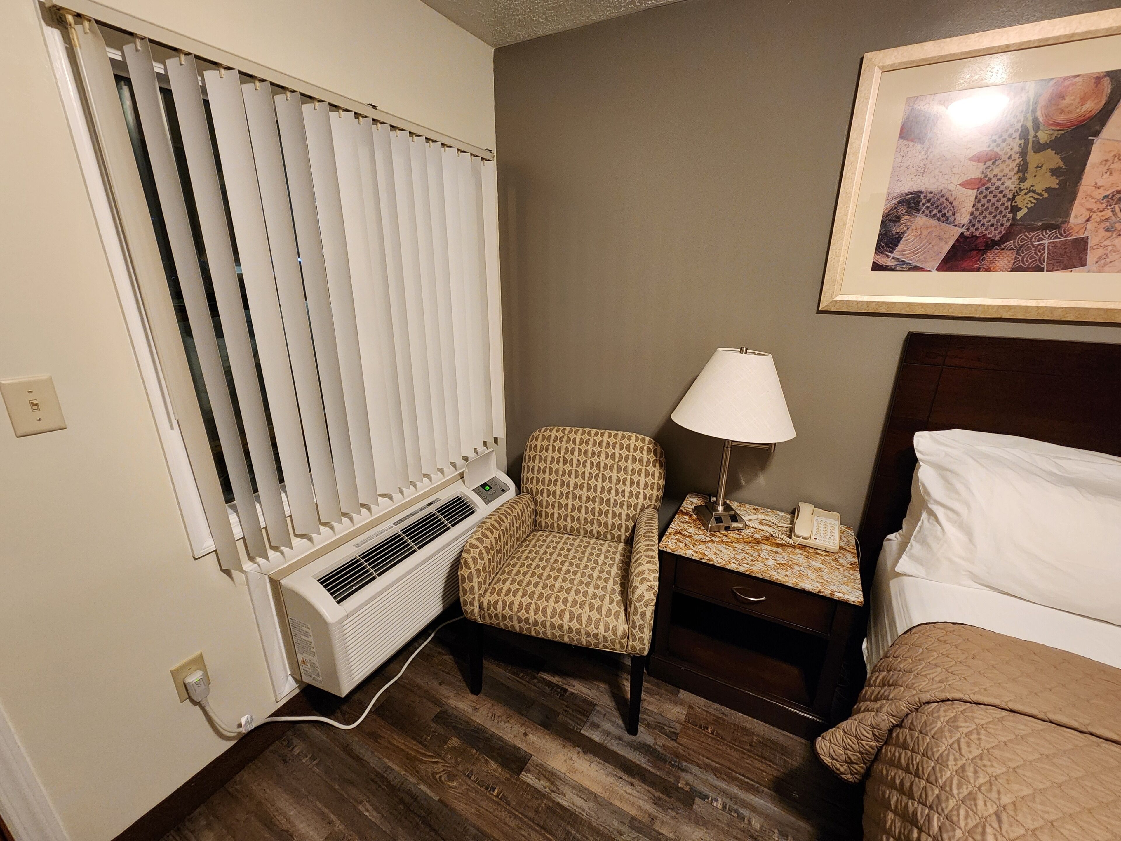 Habitación de hotel con cama, una silla, mesita de noche con lámpara, teléfono y radiador.