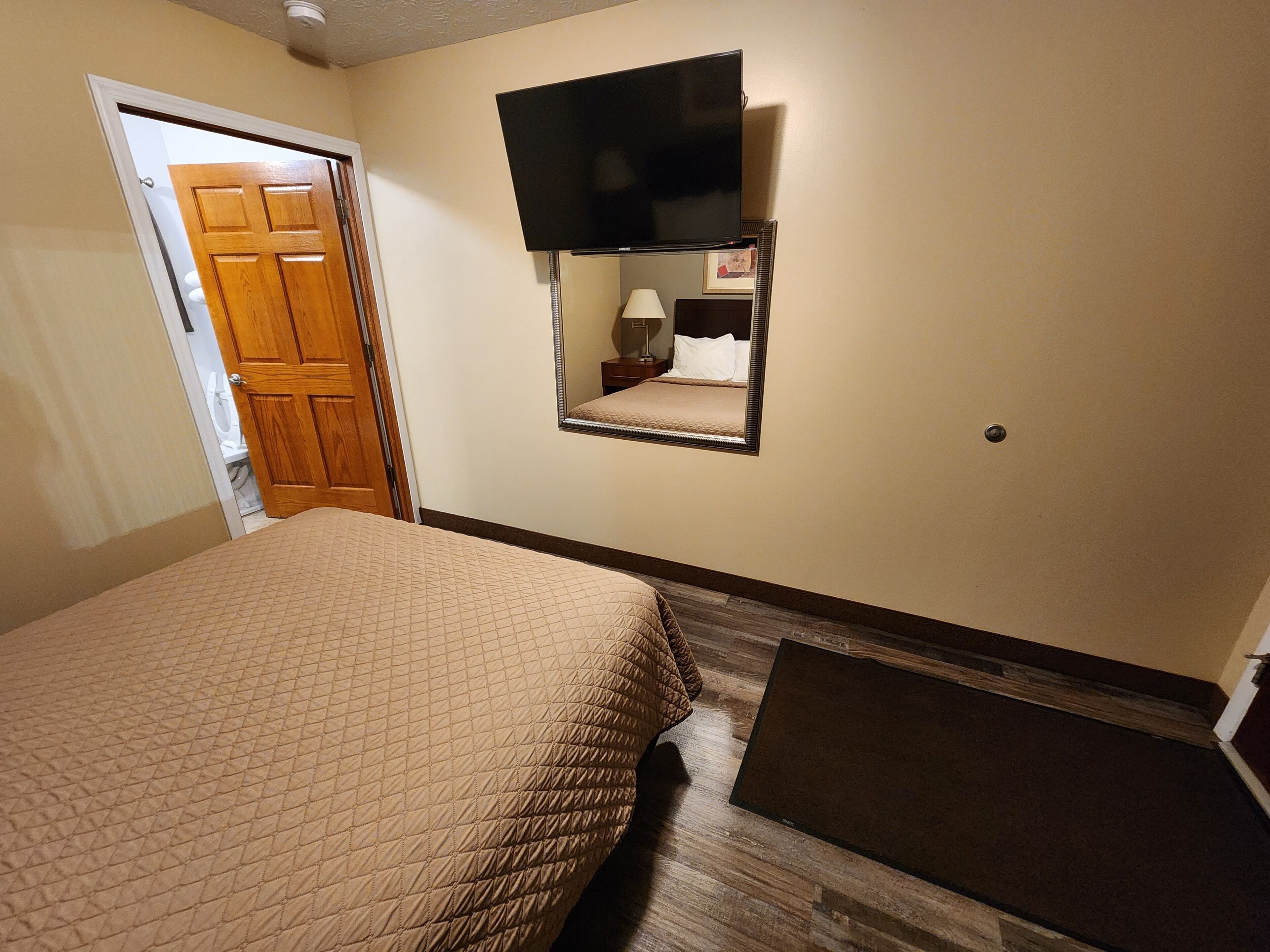 Habitación de hotel con cama, televisor montado en la pared, mesita de noche con lámpara y puerta de madera.