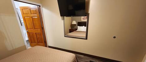 Habitación de hotel con cama, televisor montado en la pared, mesita de noche con lámpara y puerta de madera.