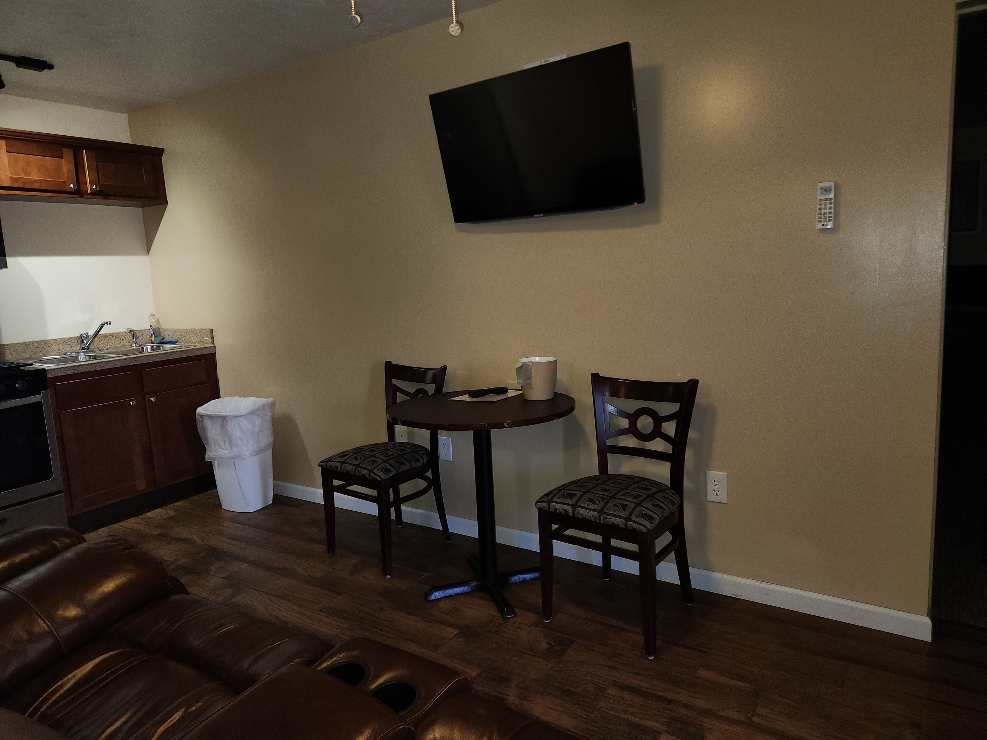 A living room with a brown leather couch, a small round table with two chairs, a TV mounted on the wall, and a kitchen area with wooden cabinets and a sink.
