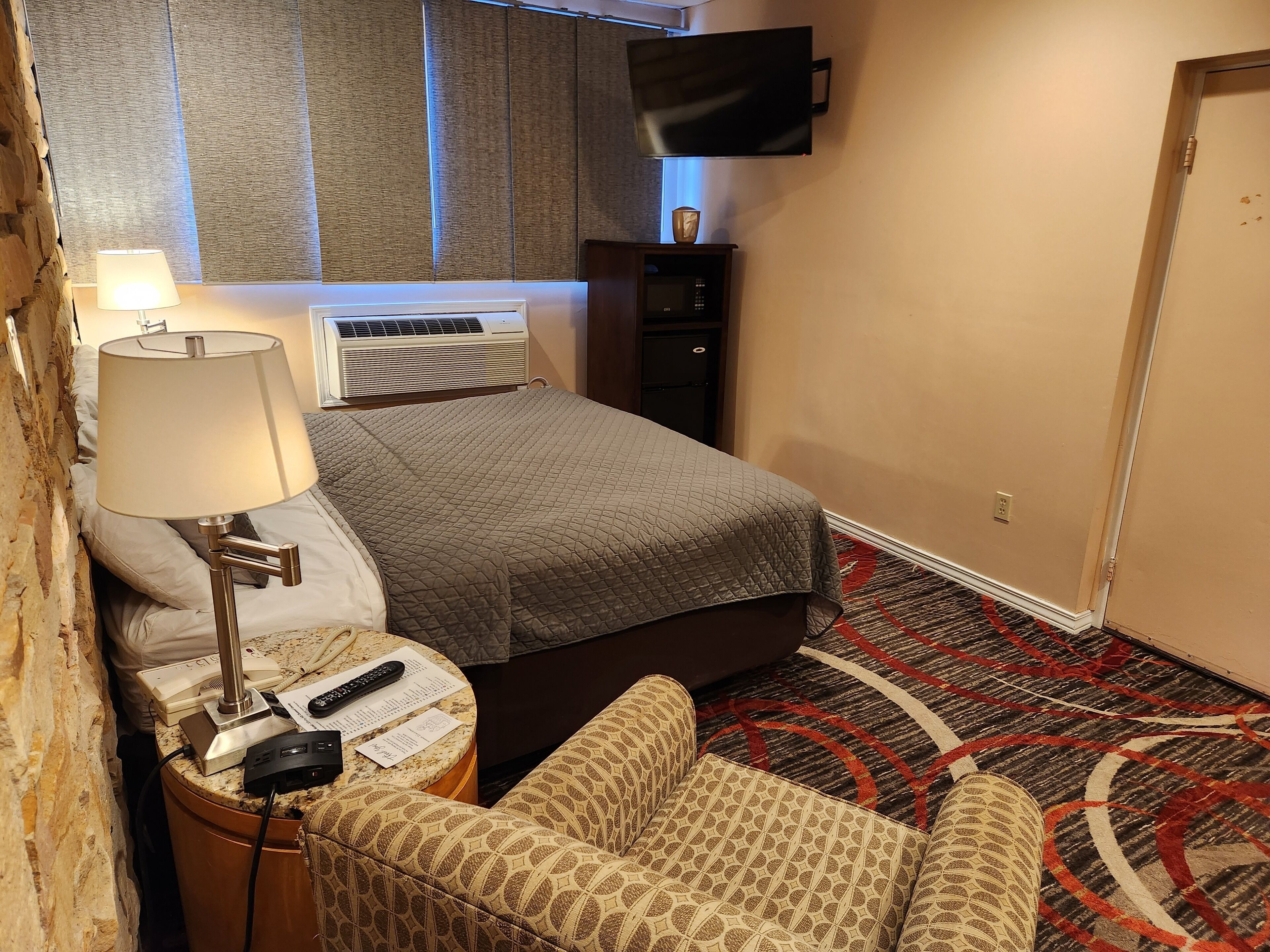 Habitación de hotel con cama, escritorio con lámpara, silla, televisor montado en la pared y alfombra estampada.