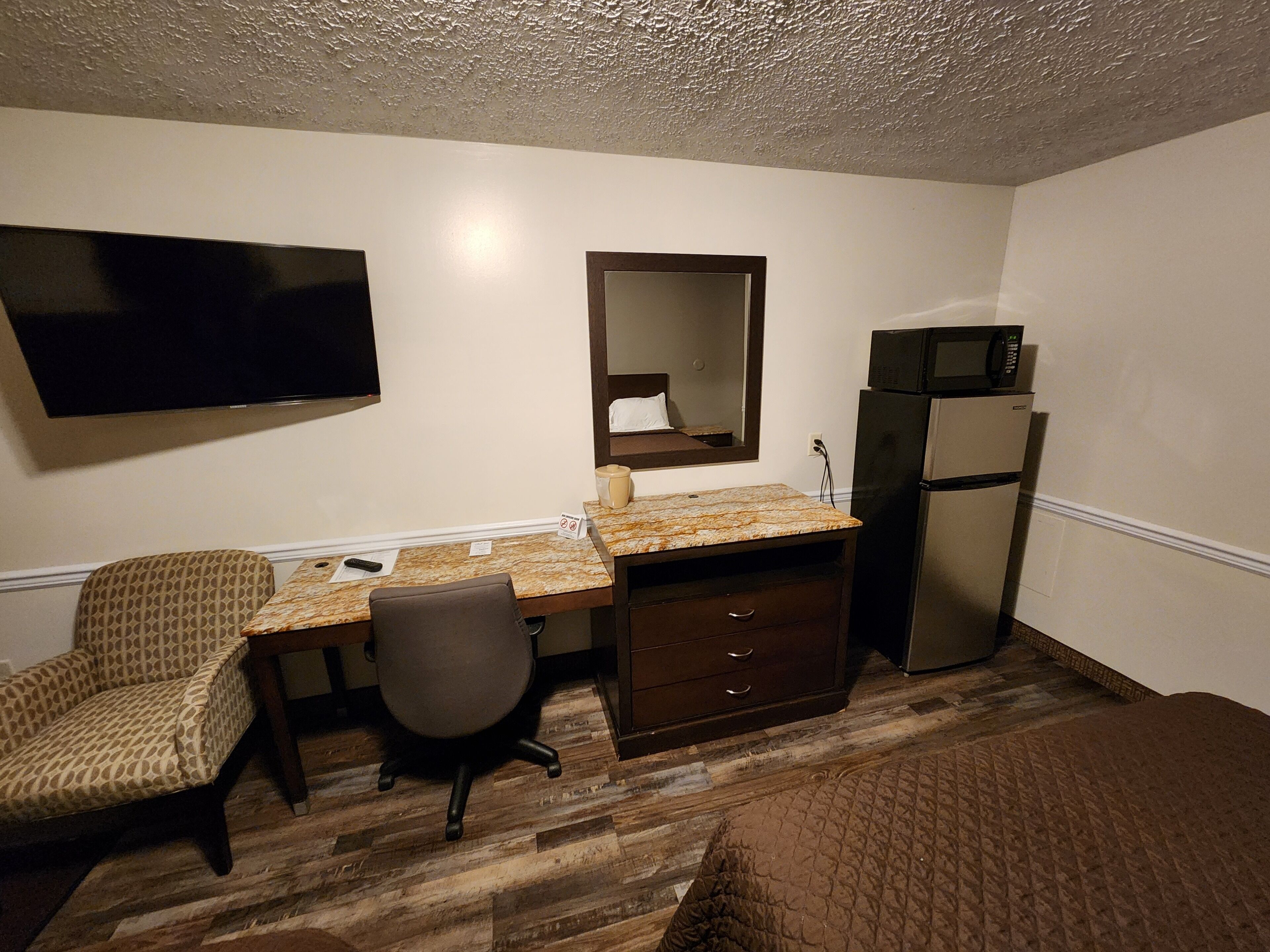 Una habitación de hotel con piso de madera, un escritorio con silla, una cama, un televisor montado en la pared y un refrigerador.