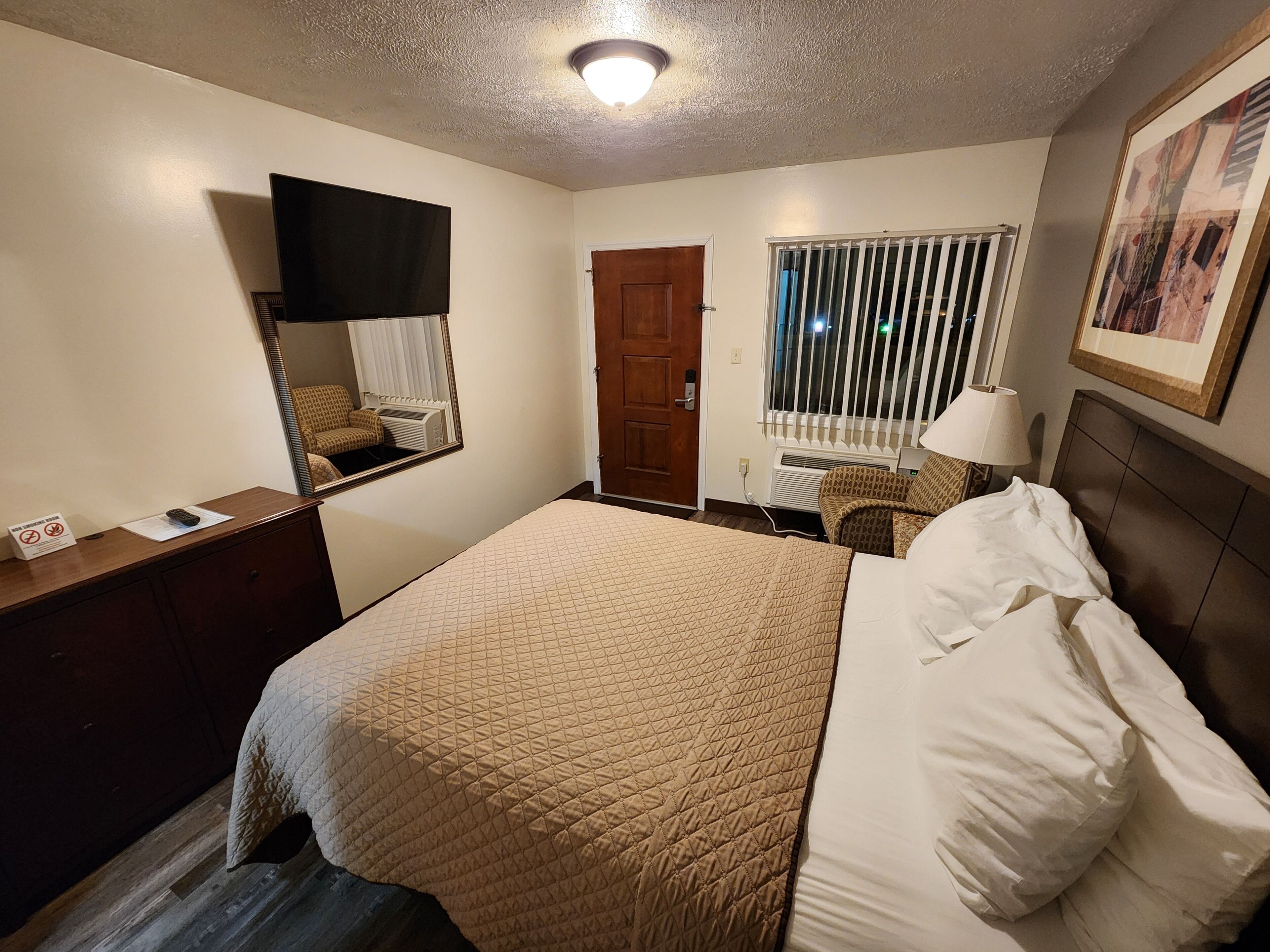 A hotel room with a bed, a desk, a chair, a window with blinds, a door, and a framed picture on the wall.