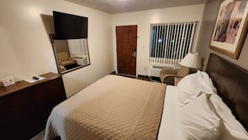 A hotel room with a bed, a desk, a chair, a window with blinds, a door, and a framed picture on the wall.