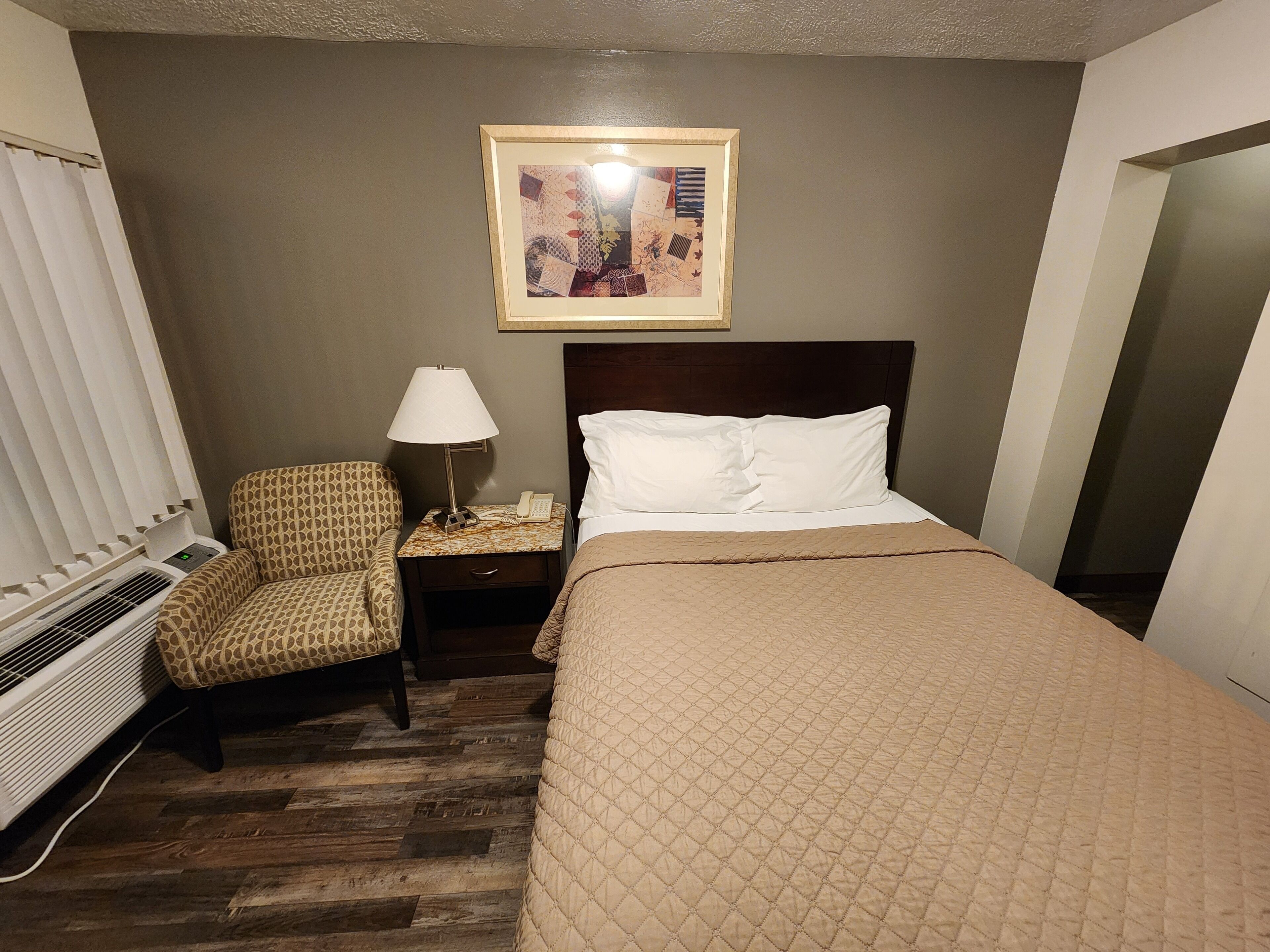 A hotel room with a bed, a chair, a nightstand with a lamp, and a framed picture on the wall.