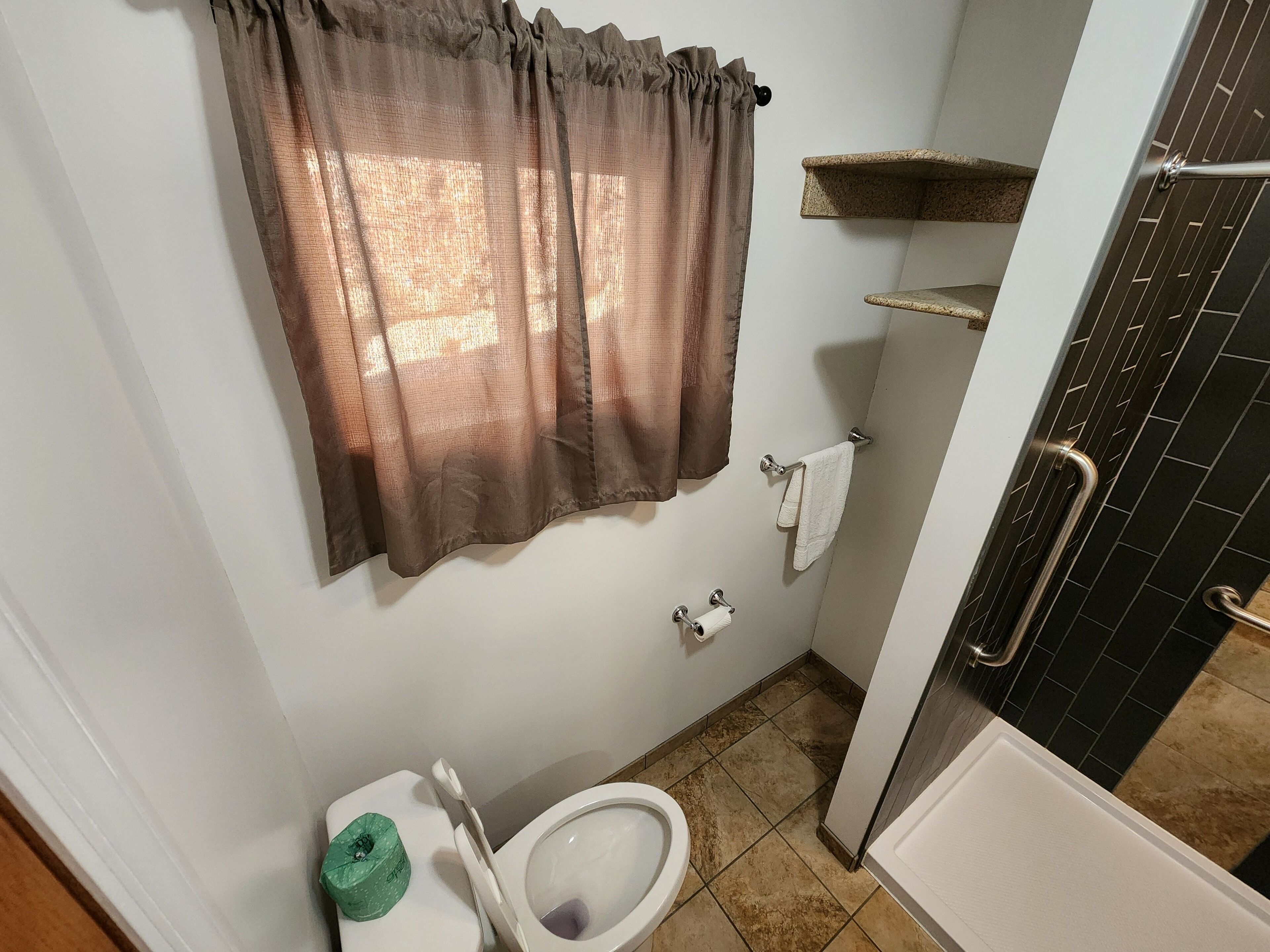 Un baño con inodoro, un dispensador de papel higiénico verde, un perchero con toalla blanca, una zona de ducha con pared de azulejos negros y una ventana con cortina marrón.