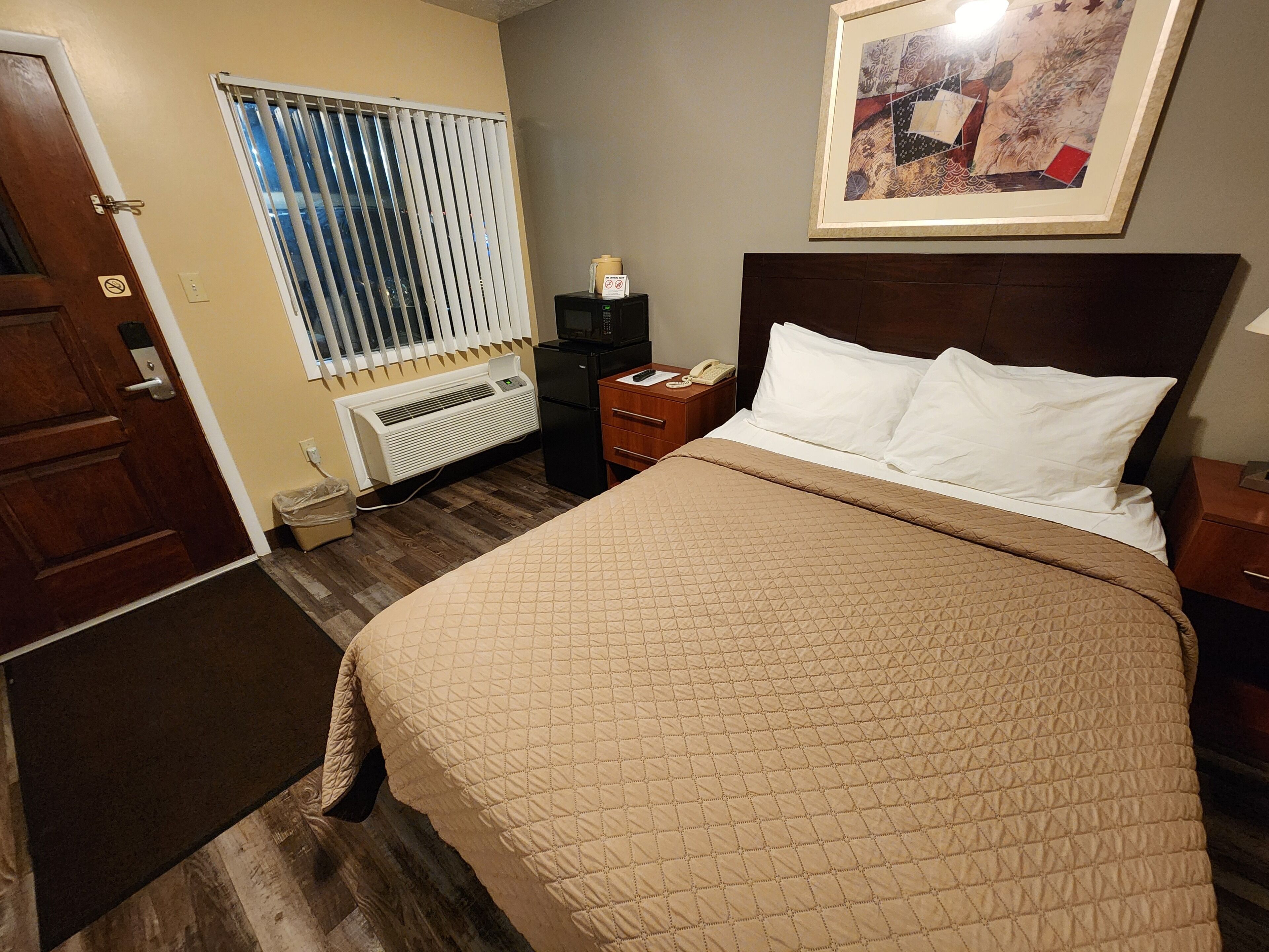 Habitación de hotel con cama, mesita de noche, teléfono, radiador y ventana con persianas.