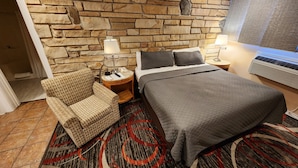A hotel room with a stone wall, a bed with a grey quilt, a patterned armchair, a small side table with a lamp, and a carpet with red and black patterns.