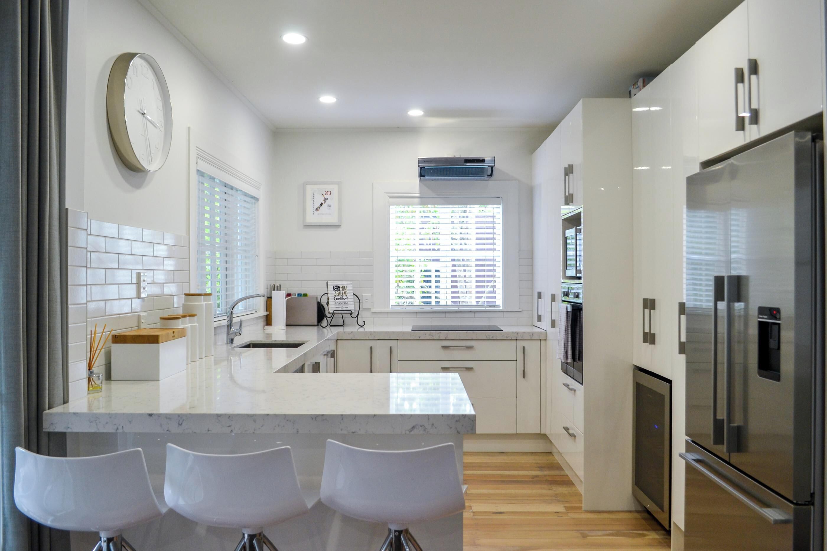 A modern kitchen with a marble island, white bar stools, stainless steel appliances, and a large window with blinds.