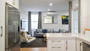 Een moderne keuken met een inbouwkoelkast, een zithoek met een stoel, een bank en een kleine tafel. Er is een raam met gordijnen, een airconditioning en een spiegel aan de muur.