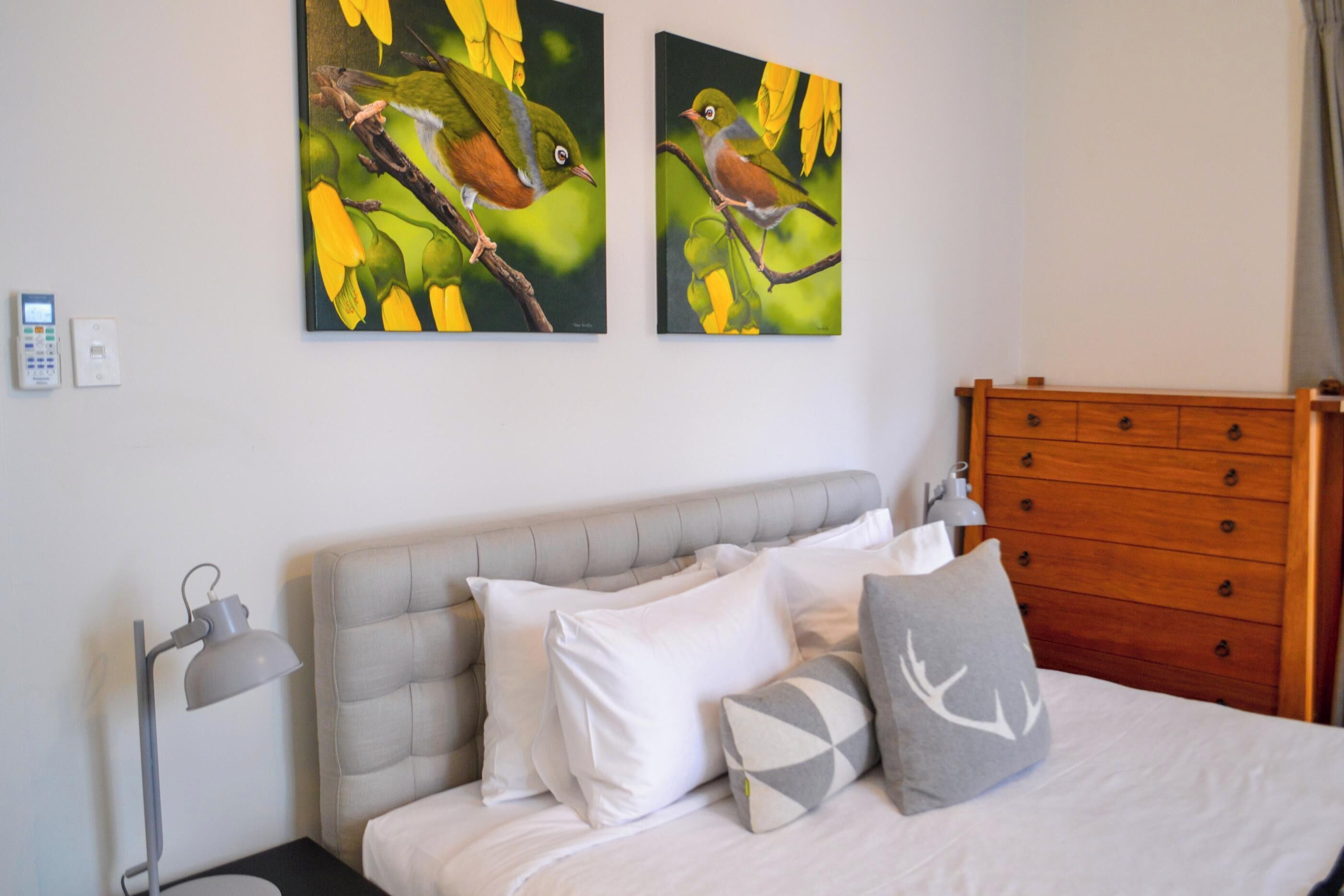 A bedroom with a bed, a wooden dresser, two wall-mounted paintings of birds, a bedside lamp, and a thermostat.