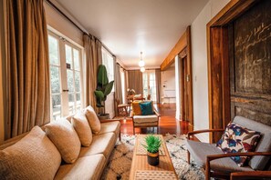 Un salon comprenant un canapé, un fauteuil et une table basse, un coin repas avec des chaises, et une grande fenêtre avec des rideaux.