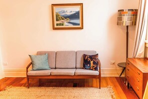 Un soggiorno con un divano, un quadro di un paesaggio, una lampada e un comò di legno.