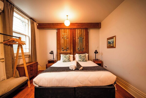 Une chambre à coucher avec un grand lit, une tête de lit en bois, deux tables de chevet, un luminaire suspendu, une fenêtre avec des rideaux et un tableau au mur.