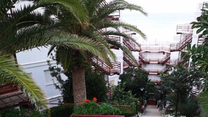 Un edificio de varios pisos con escaleras de emergencia rojas, rodeado de abundante vegetación y palmeras.