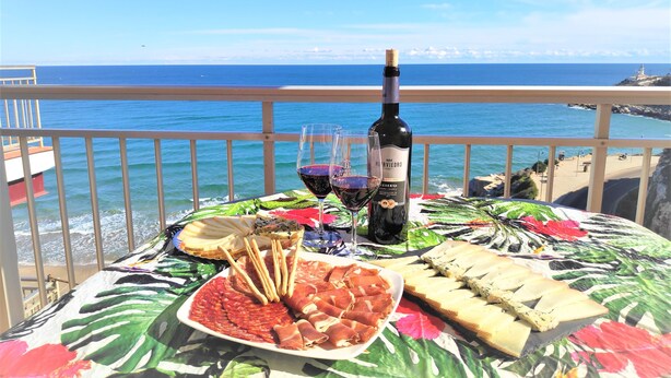 Una mesa con una botella de vino, copas y una variedad de comida, incluyendo fiambres y pan, frente a una vista panorámica del océano.