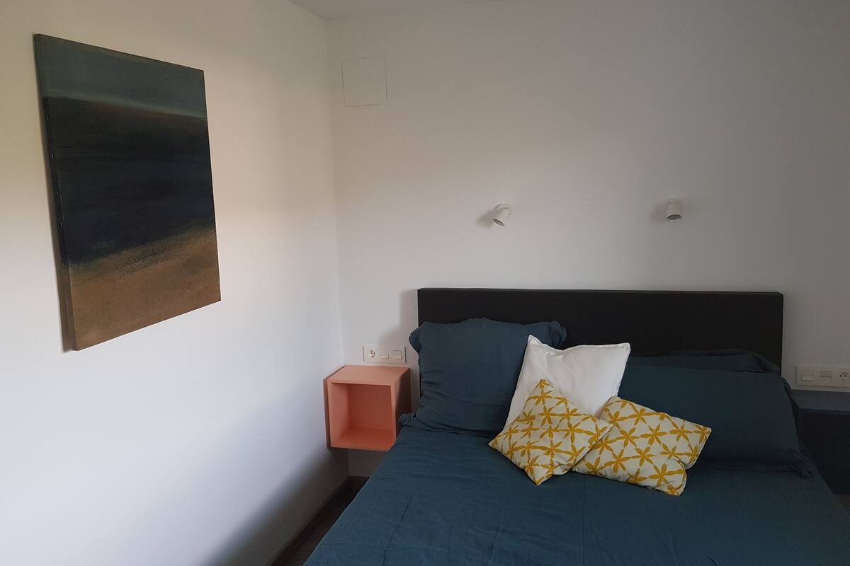 A bedroom with a bed, pillows, a nightstand, a wall-mounted light, and a painting.