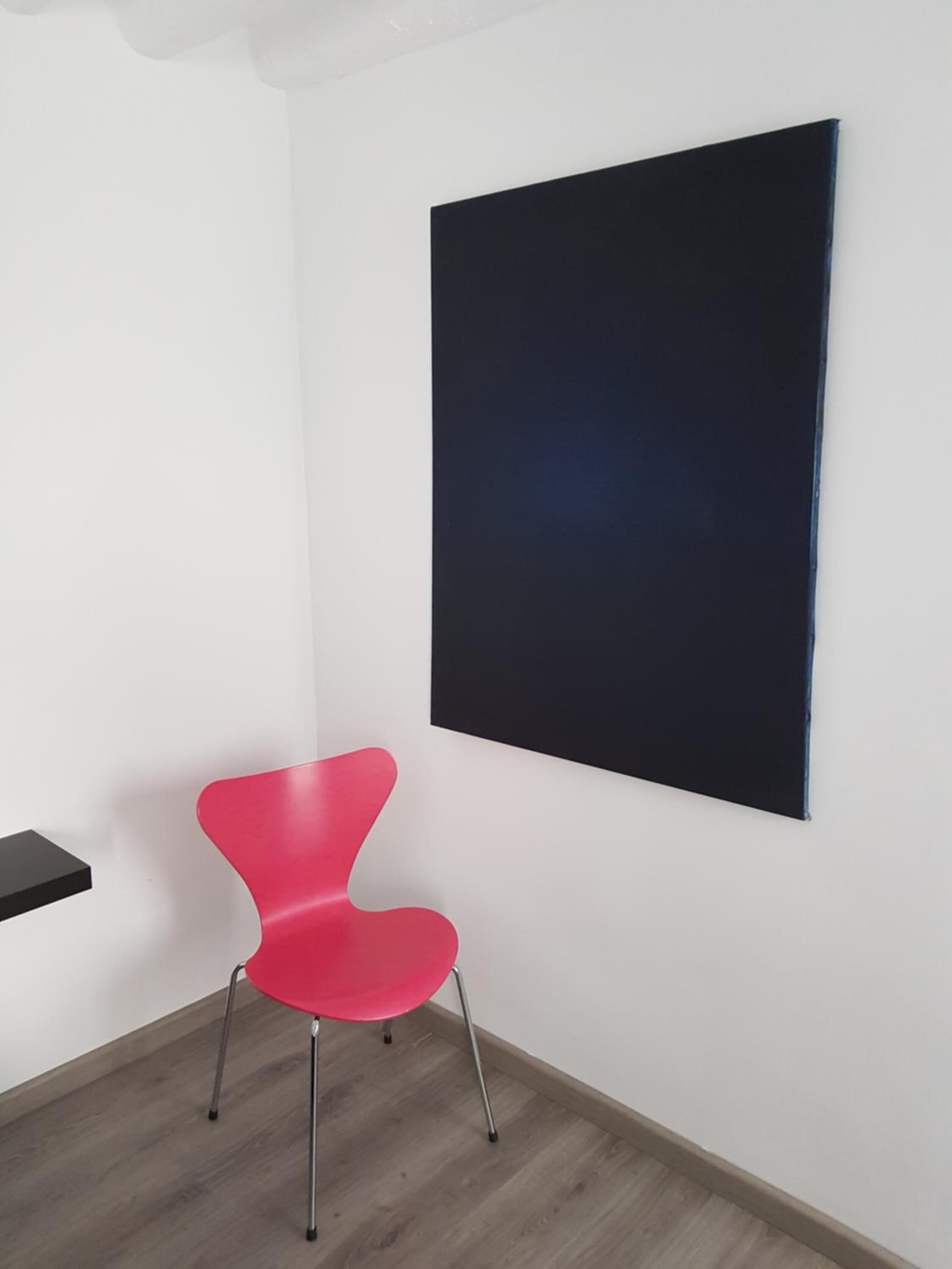 Une pièce avec une chaise rouge, une œuvre d’art noire accrochée au mur et un plancher en bois.