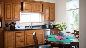 A kitchen with wooden cabinets, a black countertop, a dining table with chairs, and a window with a view of greenery. - Large House on a Hill- 15 Min Walk 2 Bart (Richmond)