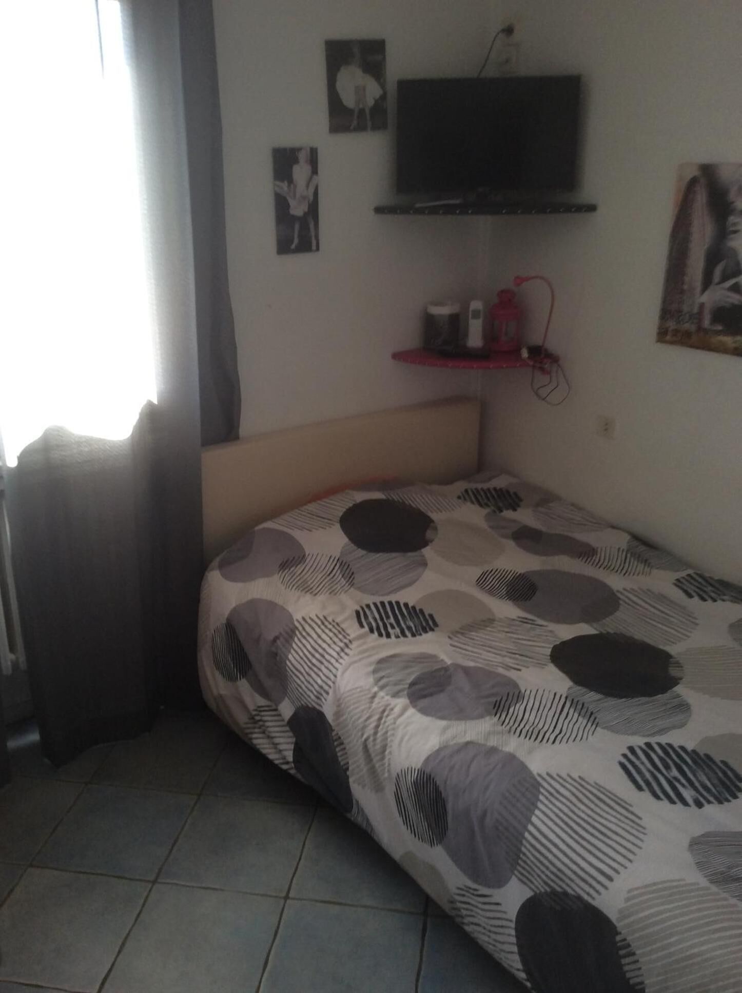 A small, single-bed room with a patterned bedspread, a mounted TV, and a small shelf with items.
