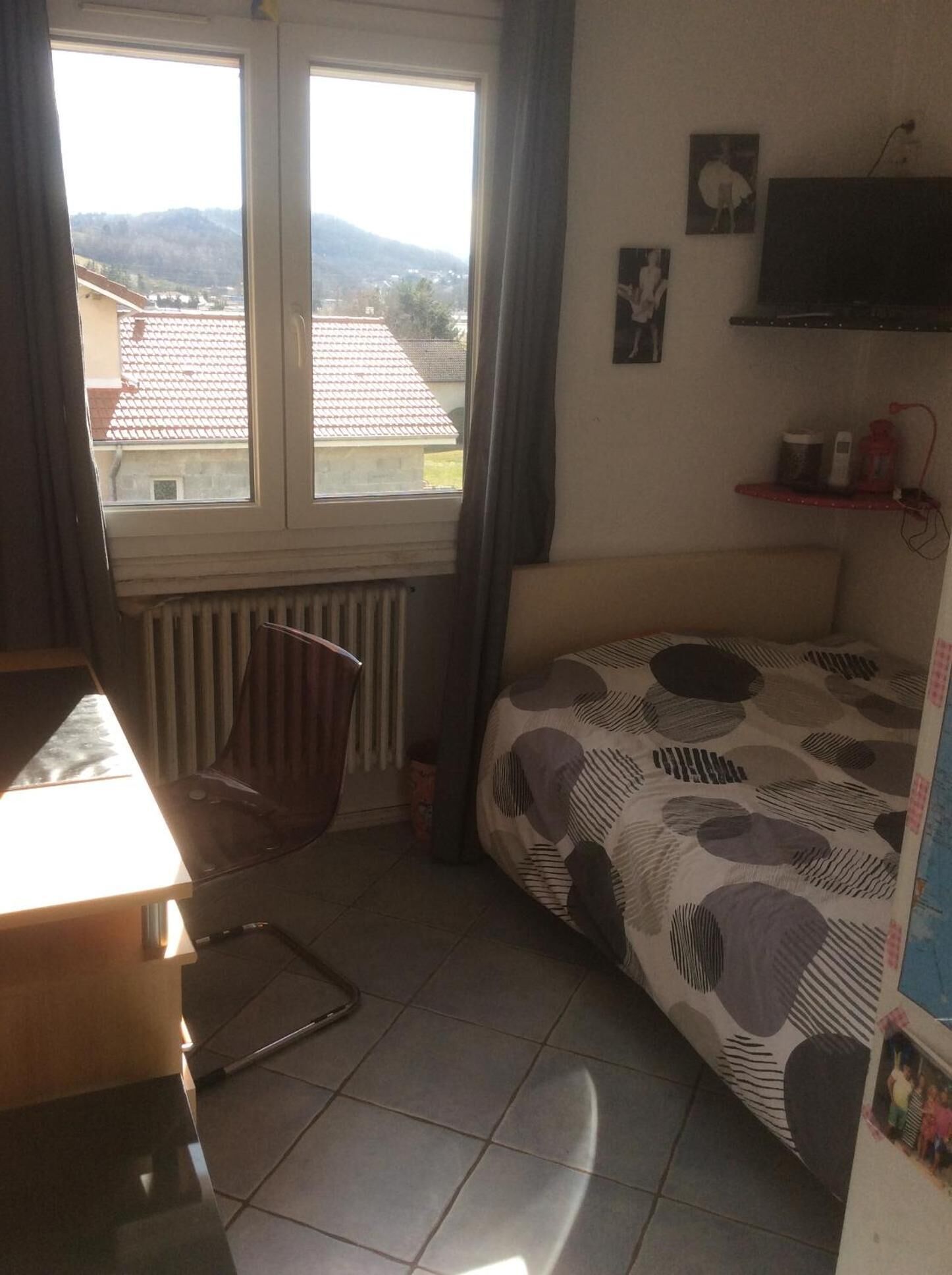 Una habitación pequeña y bien iluminada con una cama, una silla, un radiador, una ventana con vistas a edificios y colinas, y una estantería con objetos.