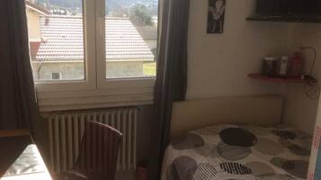 Una habitación pequeña y bien iluminada con una cama, una silla, un radiador, una ventana con vistas a edificios y colinas, y una estantería con objetos.